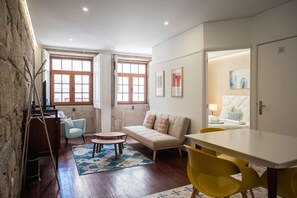 Ein gemütliches Wohnzimmer mit einer Couch, einem Couchtisch und einem Esstisch mit gelben Stühlen. Zwei Fenster mit Holzrahmen und ein Gemälde an der Wand sind zu sehen.