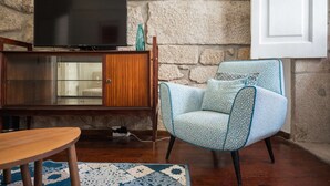 Ein Wohnzimmer mit einer Steinswand, einem Holzschrank mit Fernseher darauf, einem gemusterten Sessel und einem runden Holztisch.