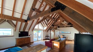 An attic-style living space with exposed wooden beams, a television, a red sofa, a kitchen area, and large windows. - The Loft at Tulka House. The Seafood capital of South Australia. (Tulka)