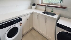 A compact kitchen with white cabinets, a built-in washing machine, and a window with a view of greenery. - The Loft at Tulka House. The Seafood capital of South Australia. (Tulka)