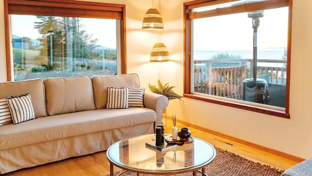 Ein Wohnzimmer mit einer Couch, einem Glastisch und zwei großen Fenstern, die einen Blick ins Freie bieten.