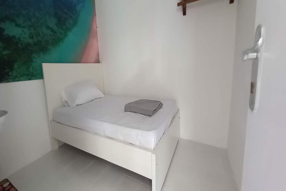 A small, white, single-bed room with a simple design, a small table, and a wall-mounted light fixture.