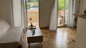 A room with a bed, a small table, and a view of a balcony with a chair and a potted plant.
