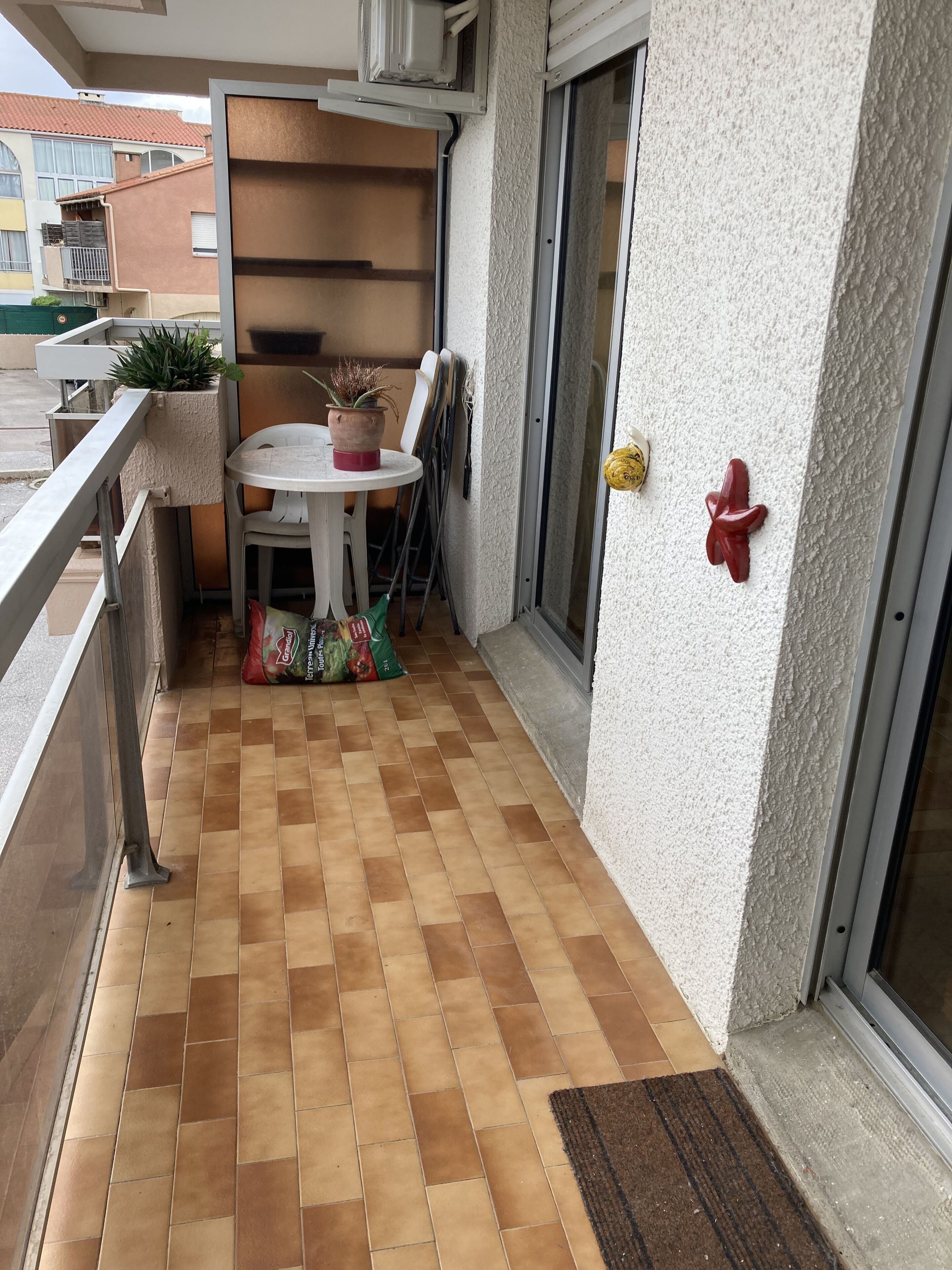 A small balcony with a tiled floor, a table with chairs, a potted plant, and a door with a doormat.