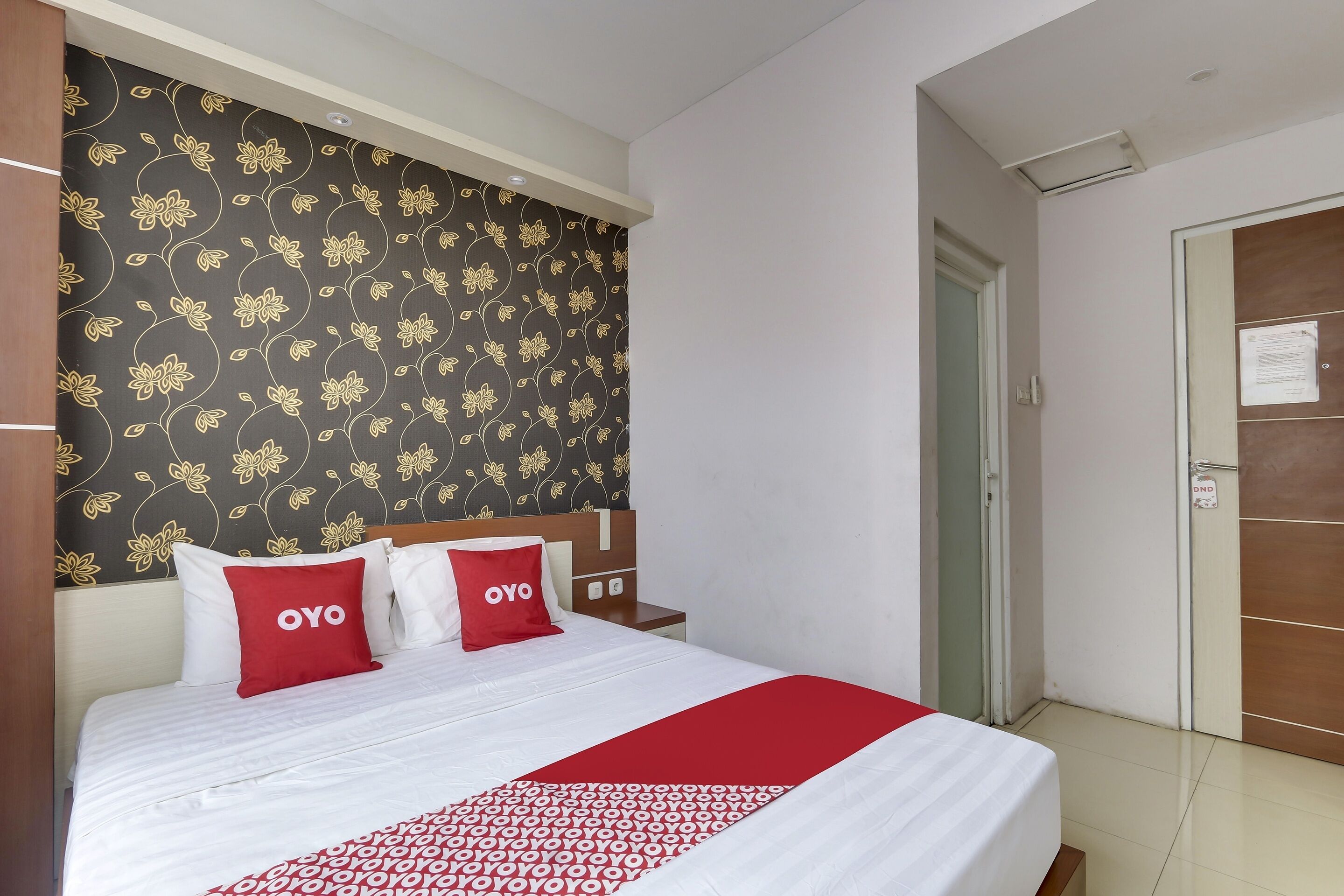 Una habitación de hotel con una cama, dos almohadas rojas con la marca 'OYO', un papel tapiz estampado y un armario de madera.