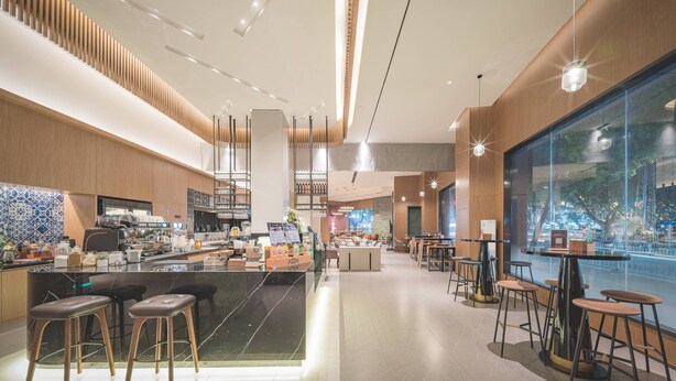 A modern cafe interior with a central bar area, seating arrangements, and a large window offering a view of the outside. - UrCove by Hyatt Nanjing Downtown (Nanjing)