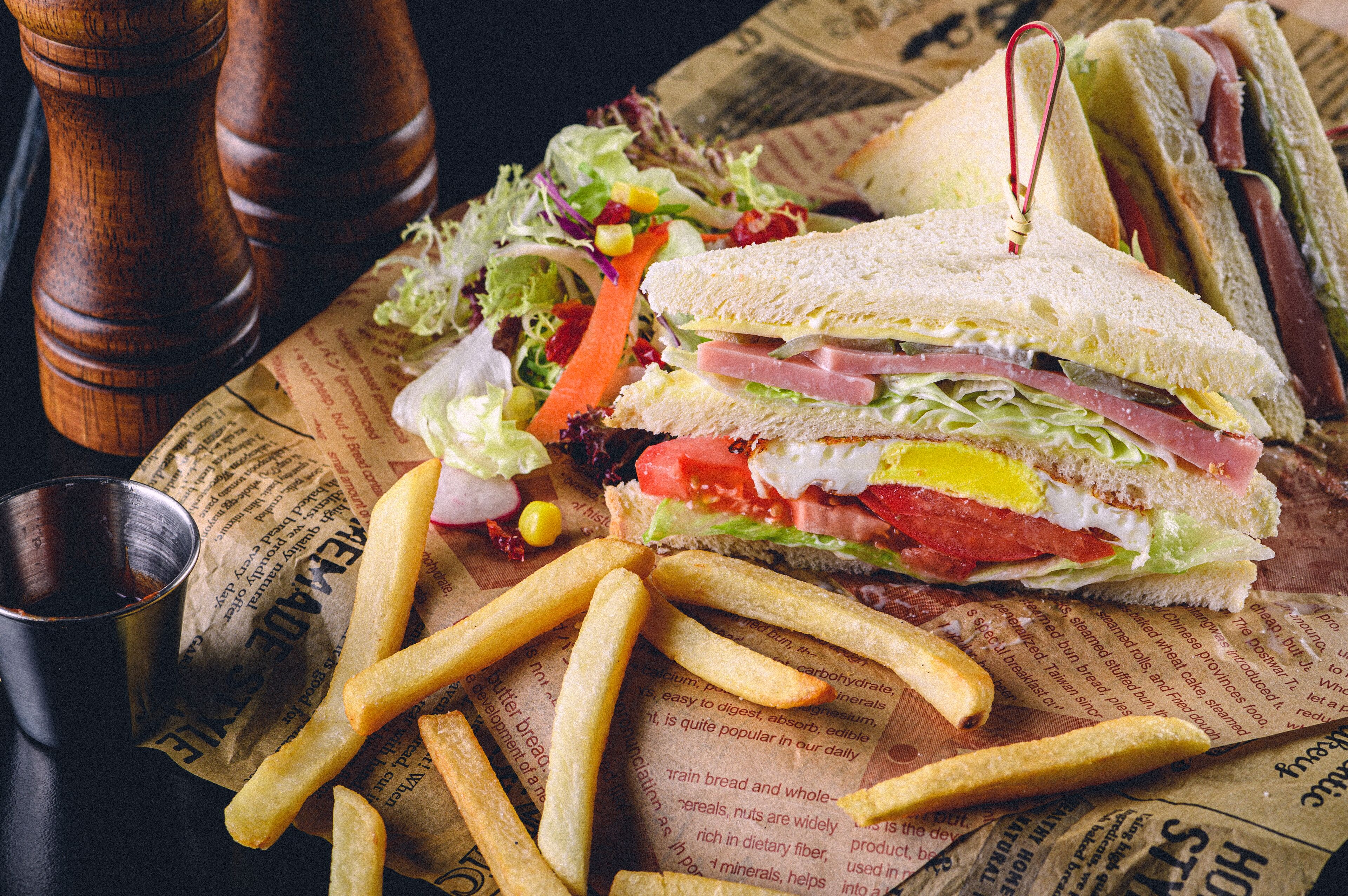 A sandwich with lettuce, tomato, and other ingredients, accompanied by a side of French fries on a newspaper.
