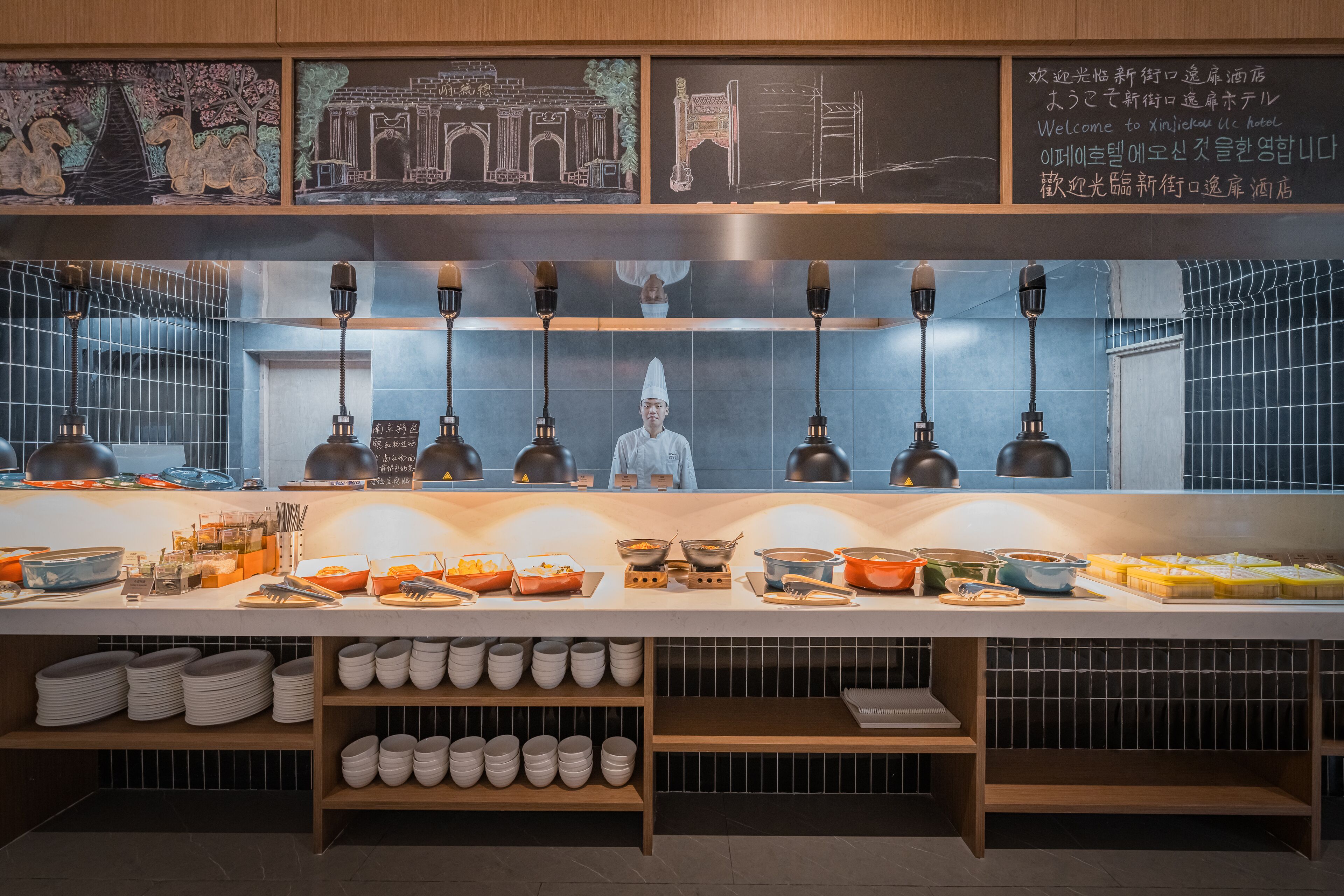 Montaje de bufé con diferentes alimentos y utensilios, un chef al fondo y un letrero con texto en un idioma extranjero.