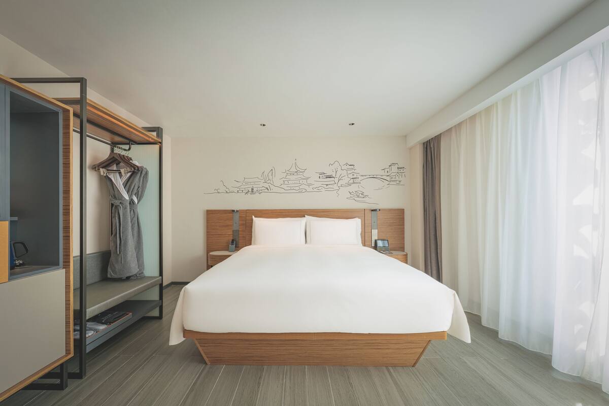 A modern hotel room with a large bed, wooden headboard, a wardrobe with hanging clothes, and a wall mural.
