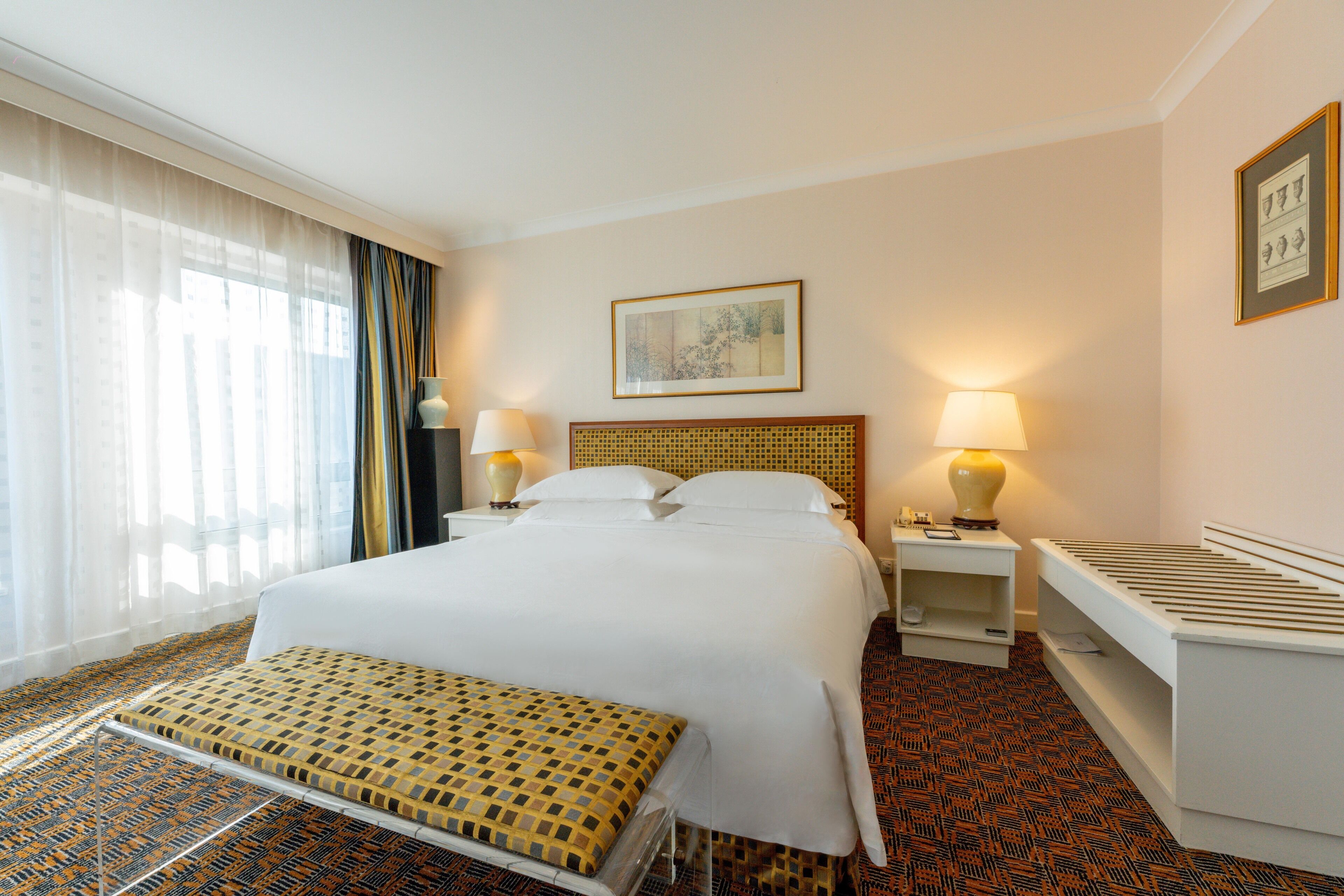 A hotel room with a large bed, two bedside tables, a bench, and a window with curtains.