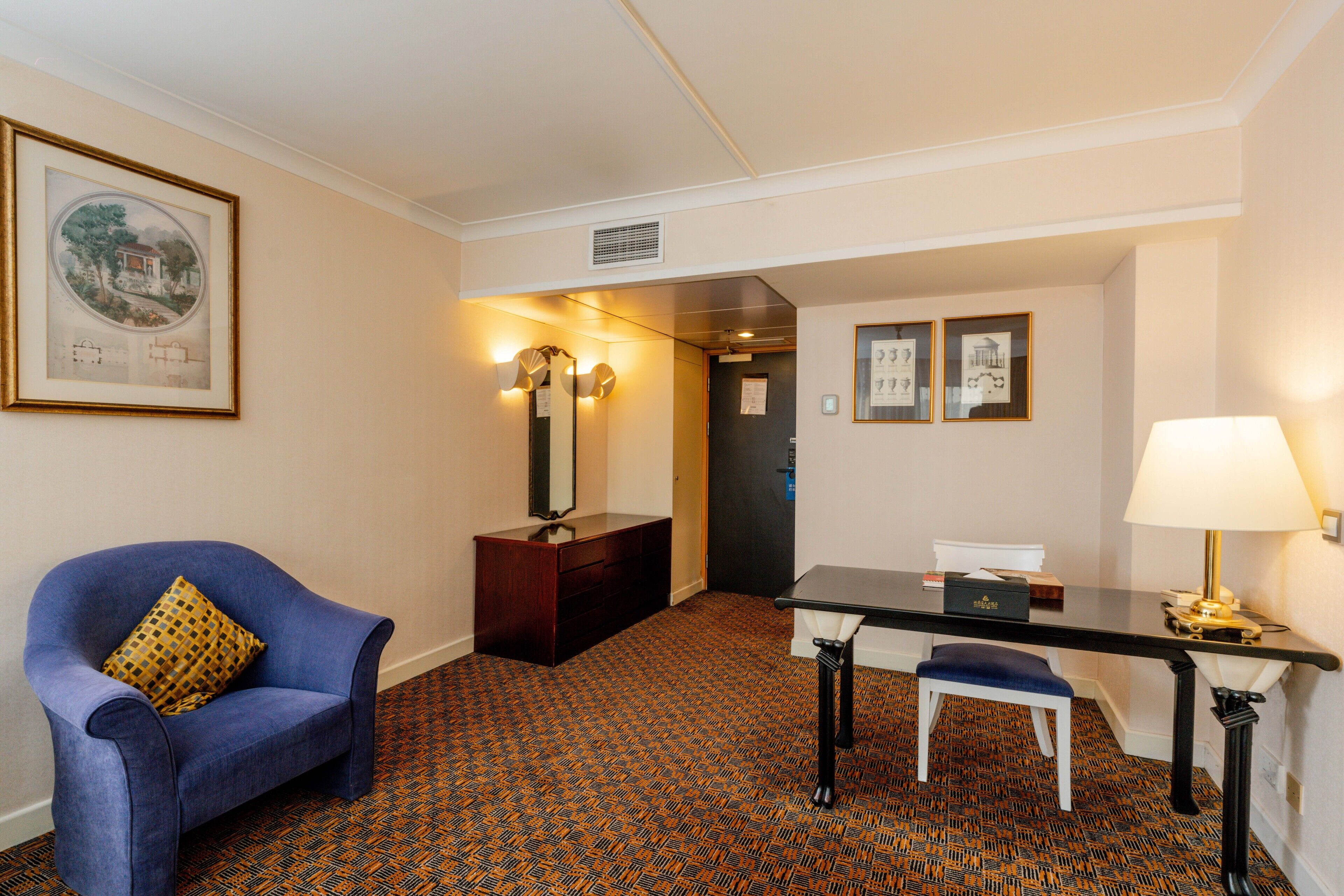 A hotel room with a blue armchair, a small table with a lamp, a desk, and a patterned carpet.