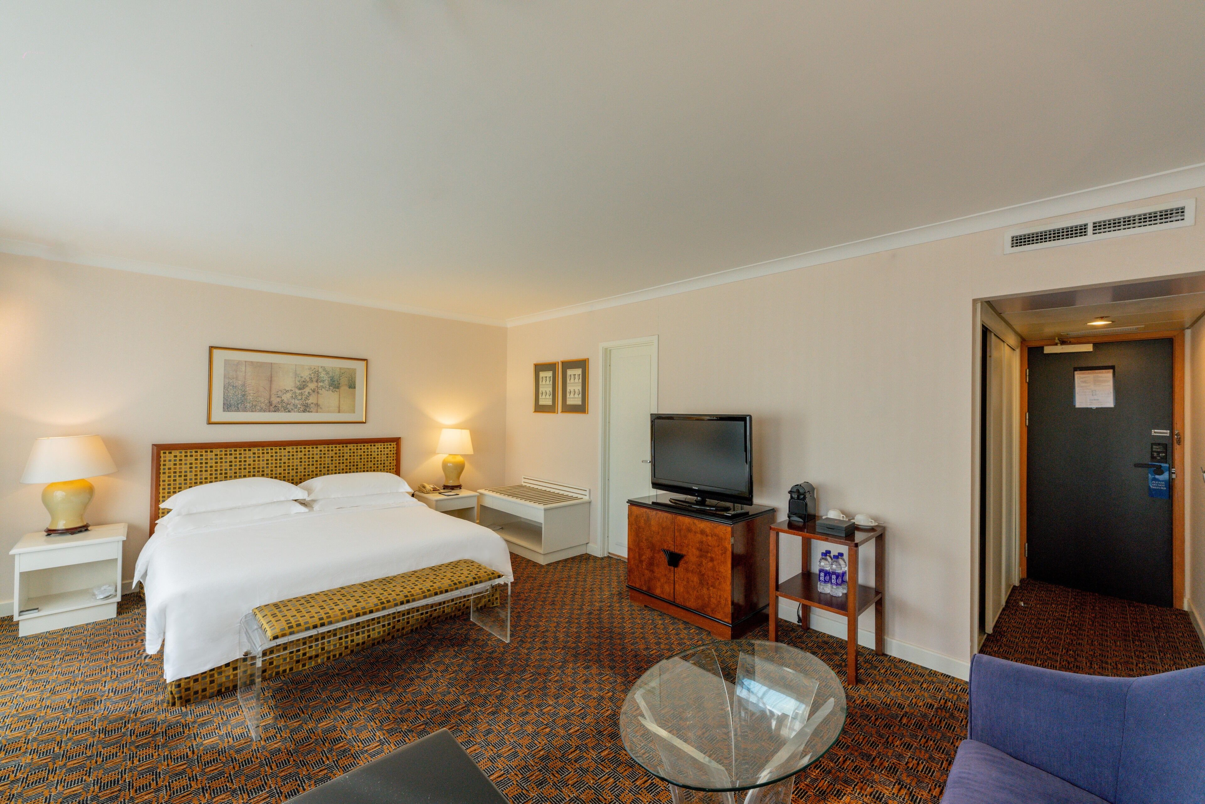 Habitación de hotel con cama, televisión, mesa de vidrio, banca, un mueble pequeño y una puerta.