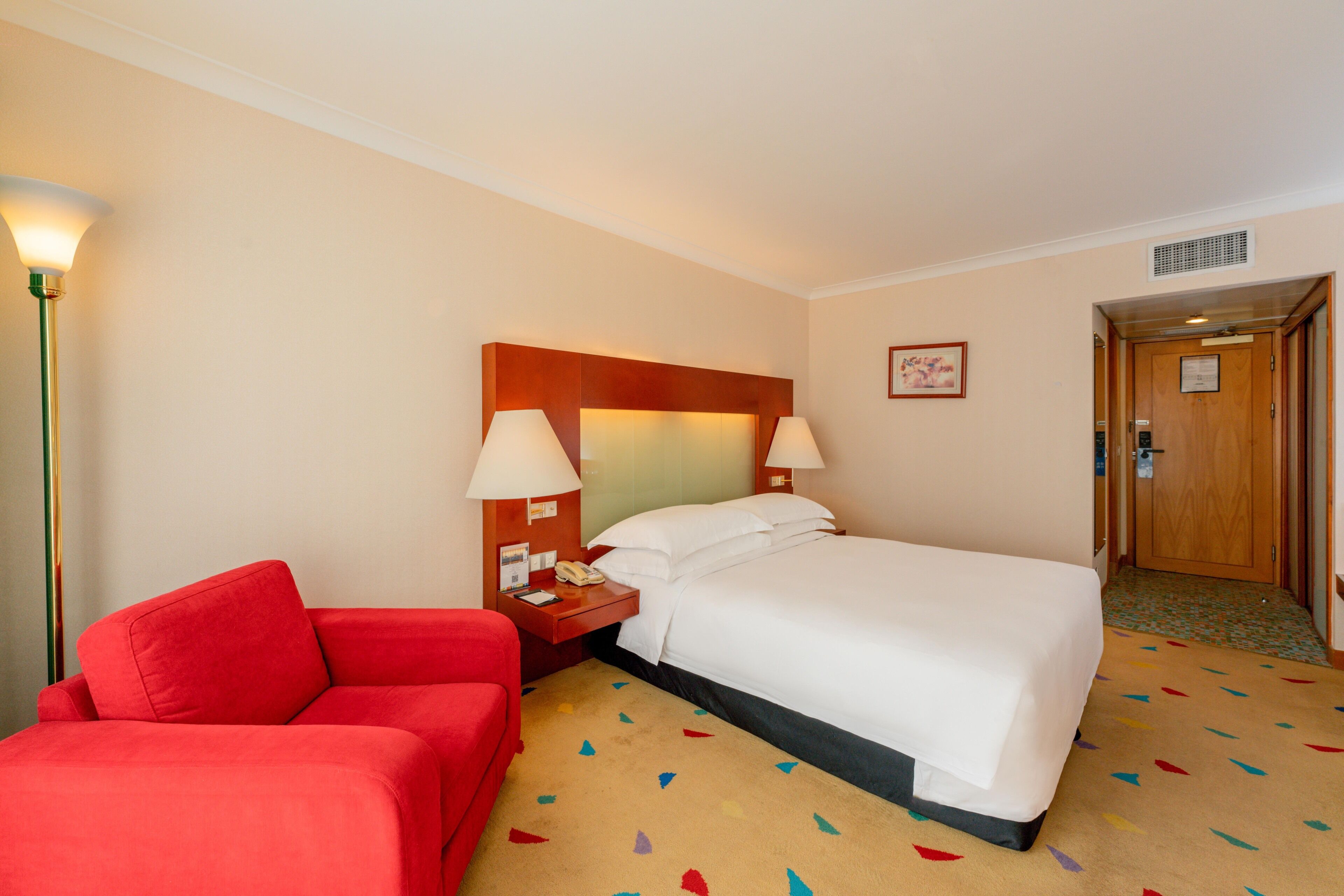 A hotel room with a bed, a red armchair, a nightstand with a lamp, and a door.