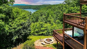 Une terrasse dotée d’un jacuzzi, d’un foyer extérieur et offrant une vue sur un lac entouré d’arbres.