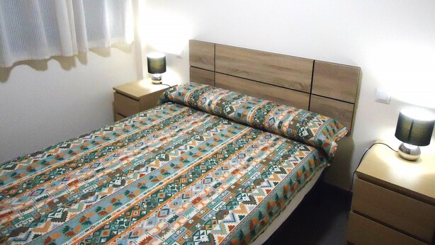 A single bed with a patterned bedspread, two bedside tables with lamps, and a window with a white curtain. - Edificio Papa Luna (Peñiscola)
