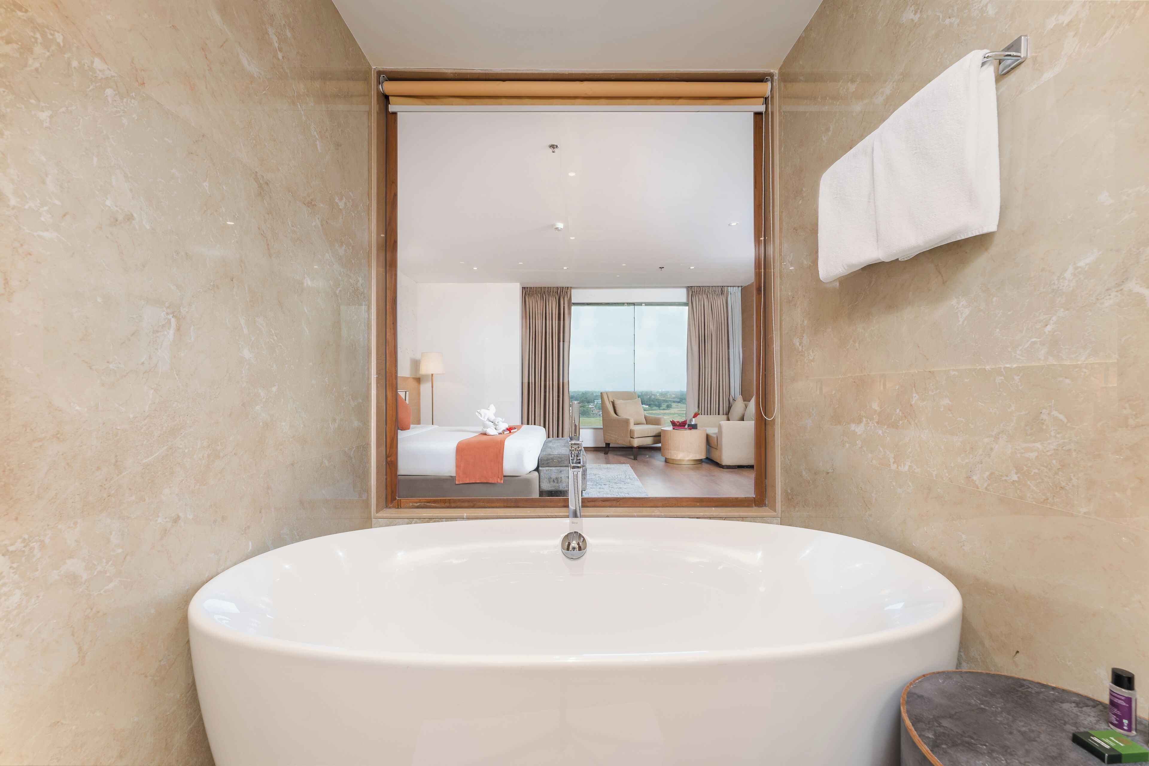 Un baño moderno con una bañera grande, un espejo que refleja un dormitorio con una cama y vistas de la ciudad.