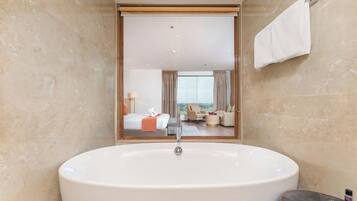 Un baño moderno con una bañera grande, un espejo que refleja un dormitorio con una cama y vistas de la ciudad.