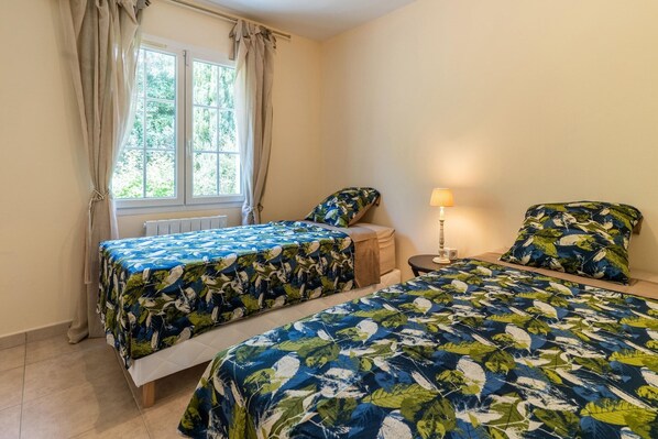 A bedroom with two beds, a window with curtains, a bedside table with a lamp, and a view of greenery outside.
