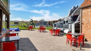 An outdoor patio area with red chairs and tables, a playground in the background, and a brick building. - Cottage on sheep farm with gelateria (Merksplas)