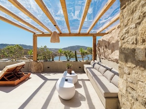 A sunlit terrace with a view of a lake, wooden beams, a hanging light fixture, a sofa, and a coffee table.