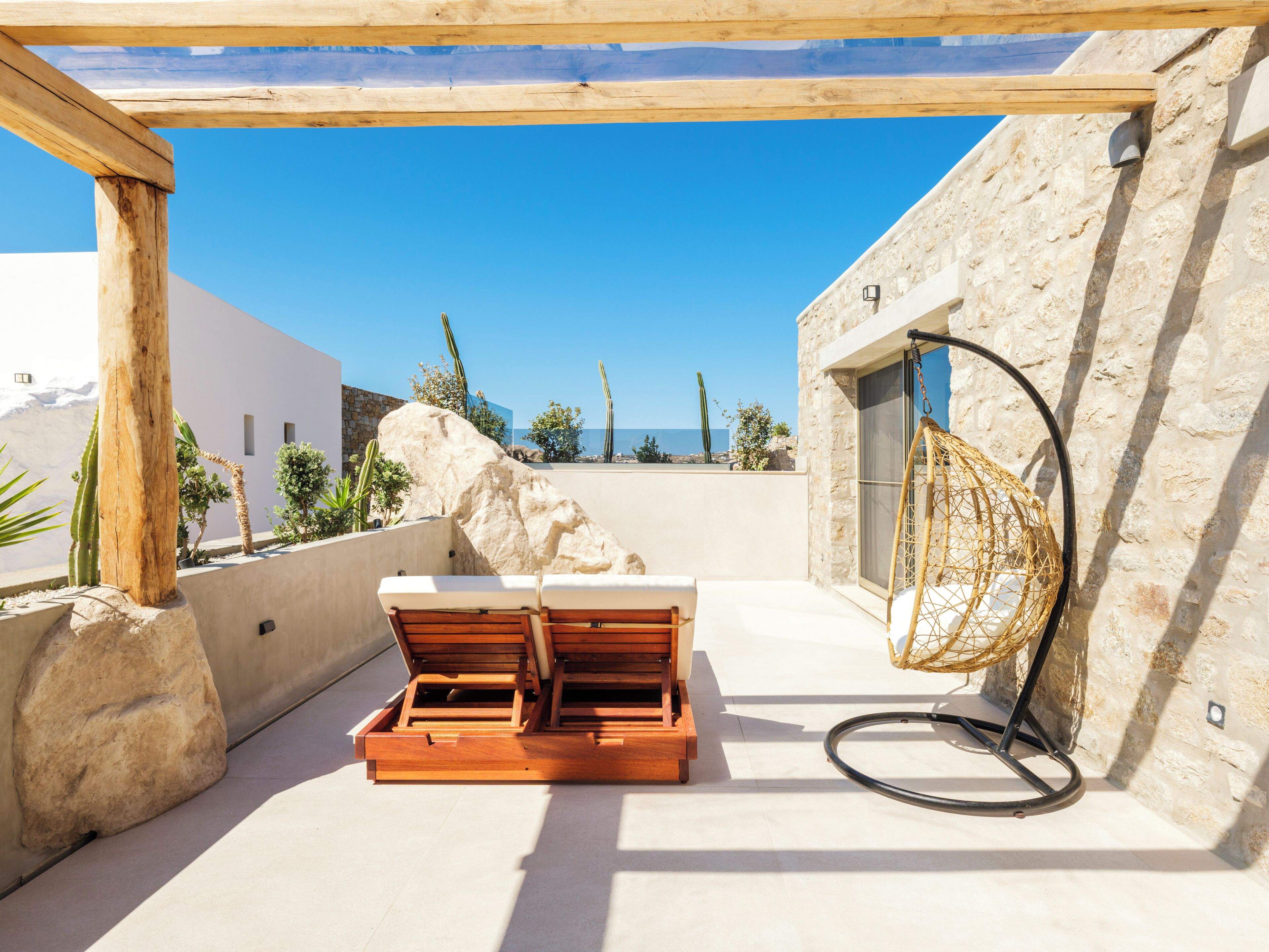 A sunlit outdoor seating area with a stone wall, a wooden pergola, and a view of the sea.