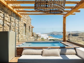 A stone exterior wall, a wooden pergola, a pool, and a view of the sea. - Nasta Suites & Villas Intentional Living Mykonos (Mykonos)