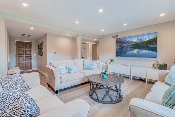 A modern living room with a beige sofa set, a round coffee table, and a large painting of a mountain lake on the wall.