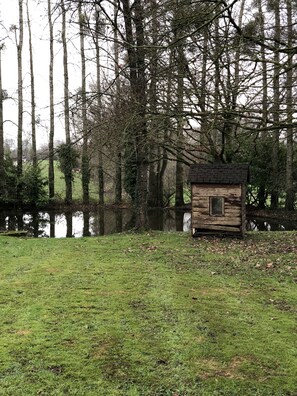Eine rustikale Holzhütte mit kleinem Fenster, umgeben von einer Wiese und einem Gewässer.