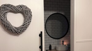 A bathroom with a white door, a heart-shaped wicker decoration, a round mirror, and a sink with a white basin.