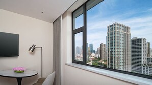 Habitación de hotel moderna con un ventanal grande que ofrece vistas a la ciudad, un televisor de pantalla plana, una mesa redonda con un florero y una lámpara de pie.