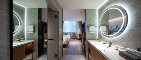 A modern hotel bathroom with two sinks, large mirrors, and a view of a bedroom with a bed and a chair.