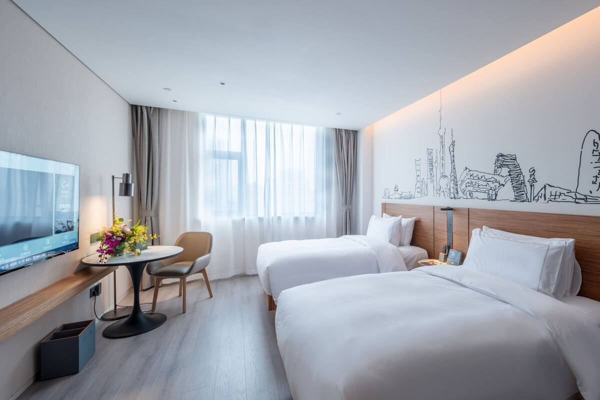 A hotel room with two beds, a TV, a small table with a chair, and a cityscape mural on the wall.