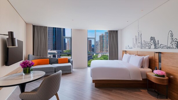 Habitación de hotel moderna con una cama grande, un sofá con cojines naranja, una mesa redonda con una silla y vistas a la ciudad a través de amplias ventanas.