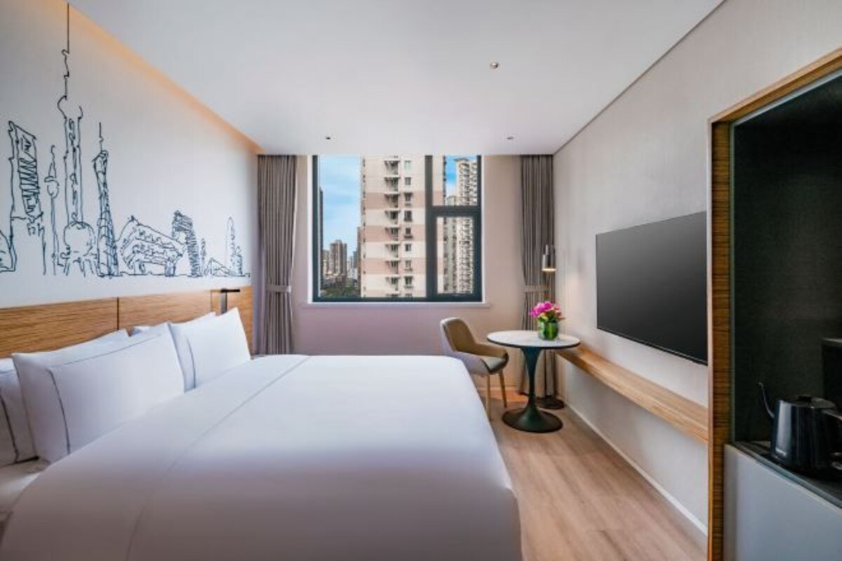 Habitación de hotel con una cama grande, una mesita con un florero, un televisor y vistas a edificios por la ventana.