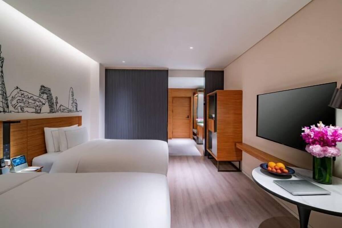 A hotel room with a bed, a desk with a laptop, a TV, and a wall with a cityscape mural.