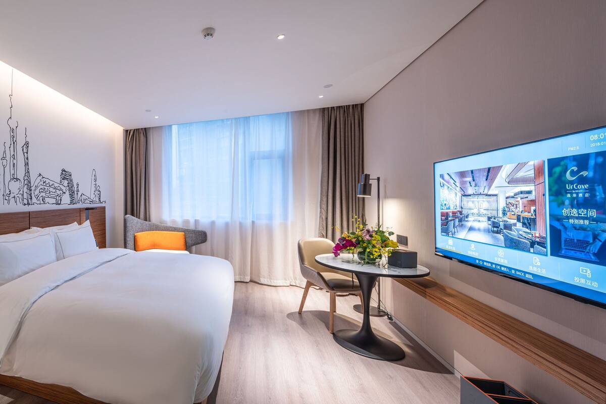 A modern hotel room with a large bed, a desk with a chair, a TV, and a cityscape mural on the wall.