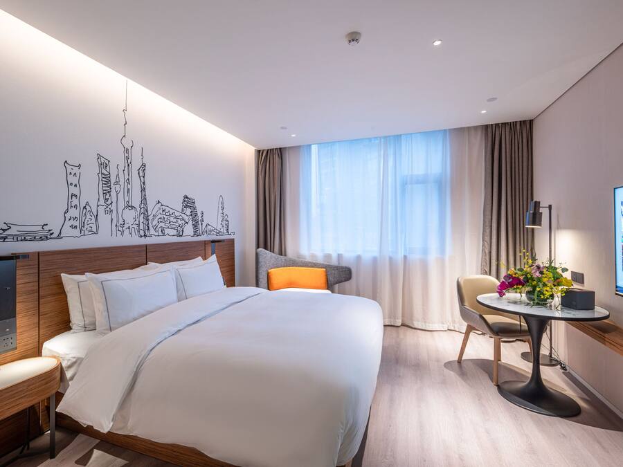 A modern hotel room with a large bed, a desk with a chair, a TV, and a cityscape mural on the wall.