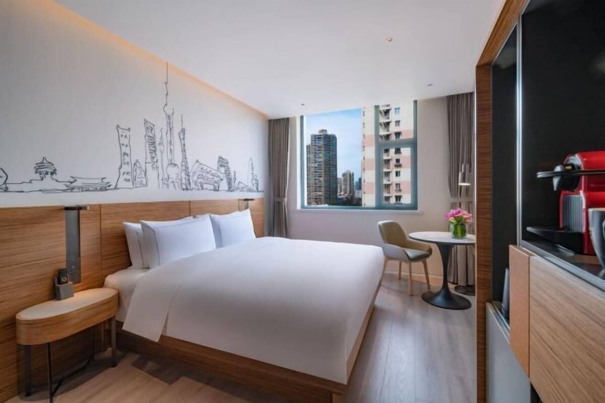 A hotel room with a large bed, a desk, a chair, a small table with a vase, and a cityscape mural on the wall.