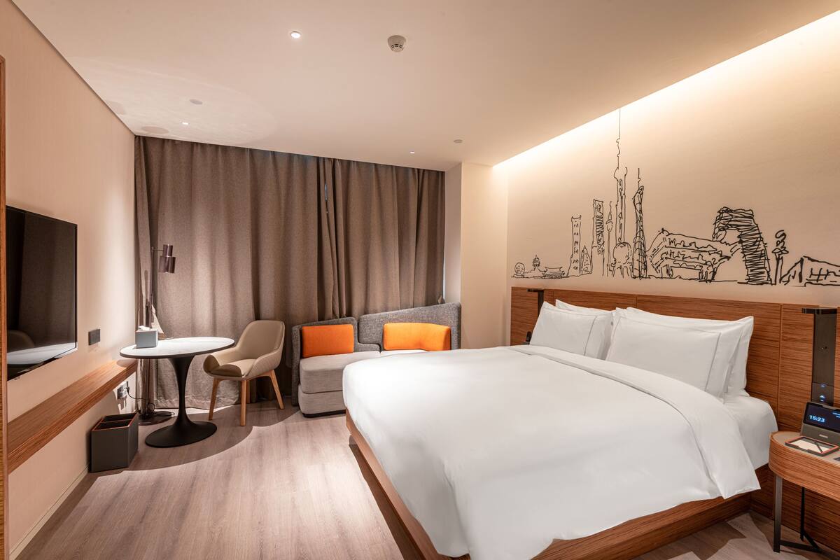 A hotel room with a bed, a desk, a chair, a small table, a TV, and a wall mural.