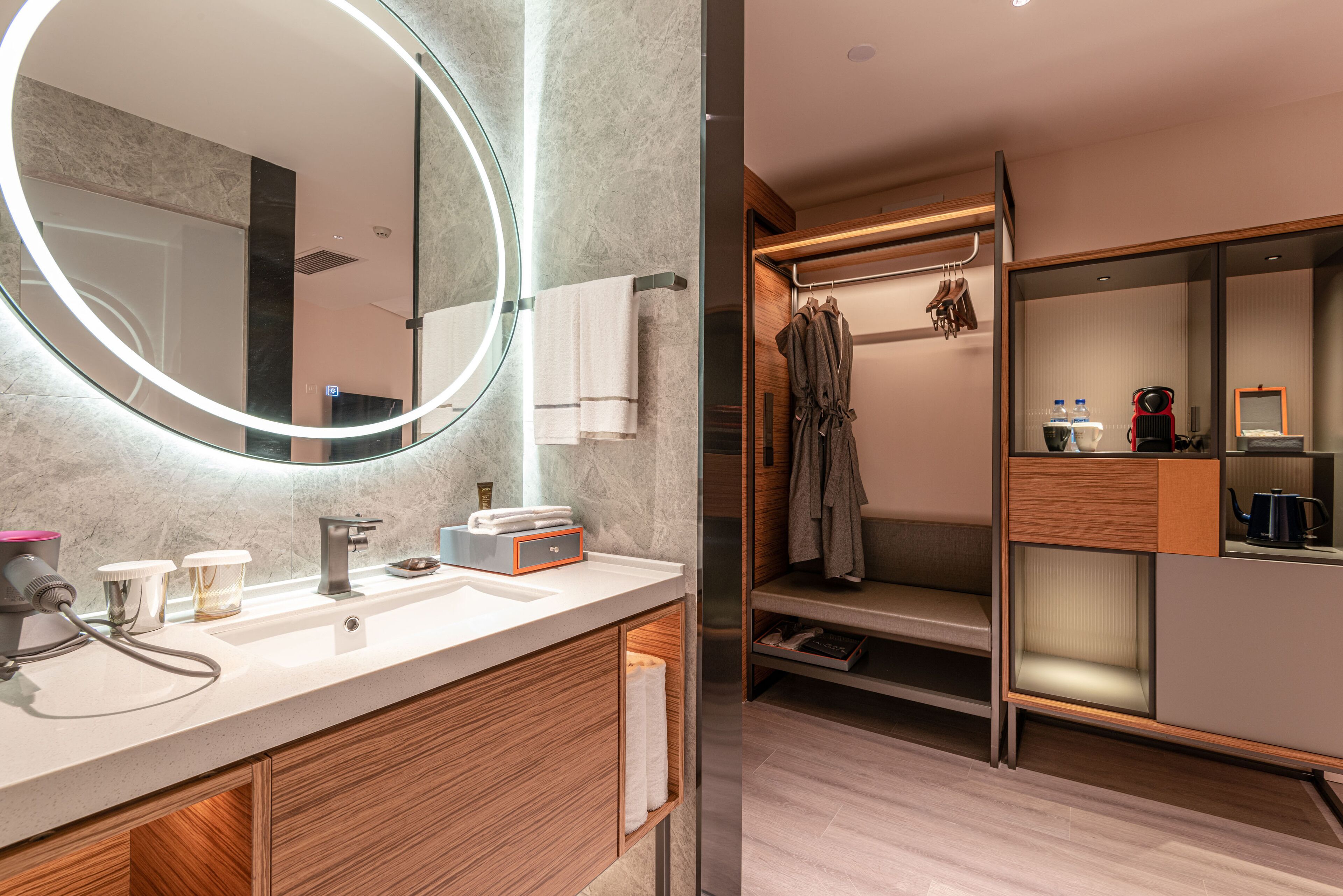 A modern bathroom with a large mirror, a sink, and a vanity. There is a robe rack with towels and a coffee machine.