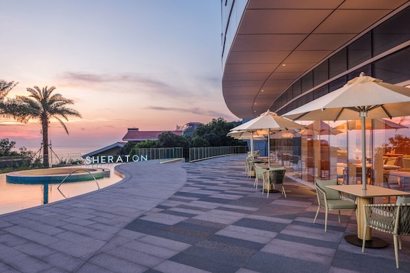 Un espace extérieur aménagé avec des parasols et des chaises, une piscine et un bâtiment sur lequel on peut lire l’inscription « SHERATON ».