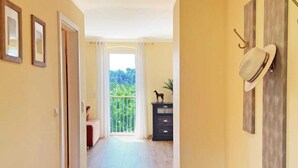 A hallway with wooden flooring, a door on the left, a wall-mounted hat rack, and a view to a balcony with a railing.