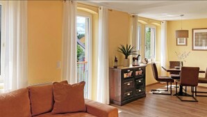 A living room with a brown sofa, a dining area with a table and chairs, a dark wooden cabinet, and a large window with curtains.