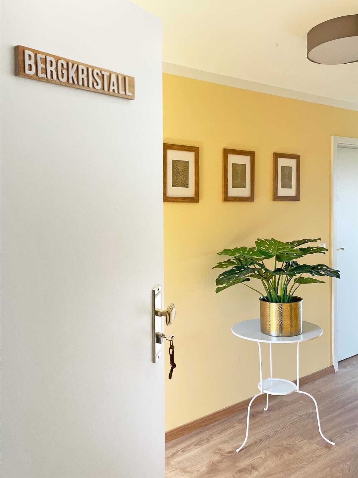 Un couloir avec une porte blanche portant l’inscription « BERGKRISTALL », une petite table blanche sur laquelle est posée une plante en pot, et trois tableaux encadrés accrochés au mur.