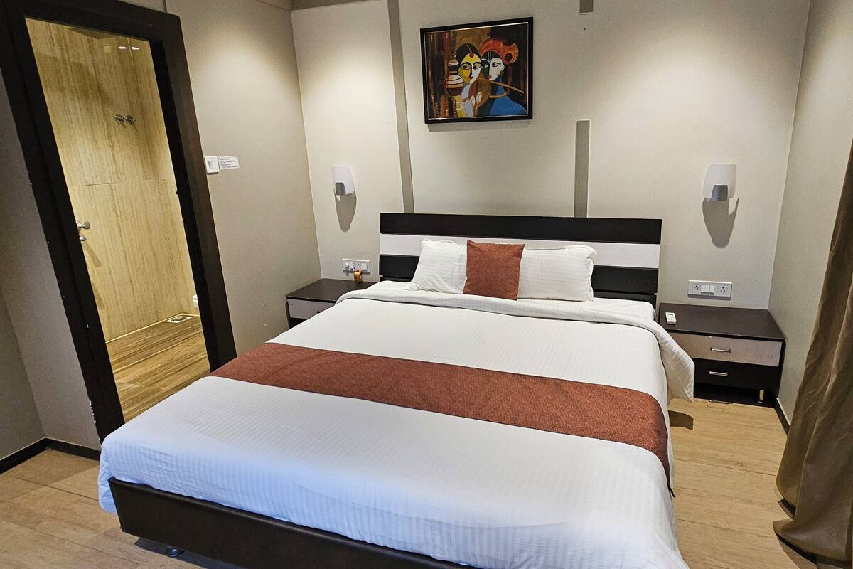 A hotel room with a large bed, a wooden door, a painting on the wall, and a nightstand with a lamp.