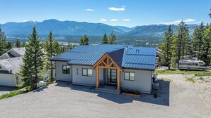 A single-story building with a blue metal roof and wooden accents, surrounded by a gravel driveway and trees, with a mountainous backdrop. - Fairmont Peak Cabin | Pet Friendly | Pool Table (Area F (Columbia Lake/Windermere Lake))