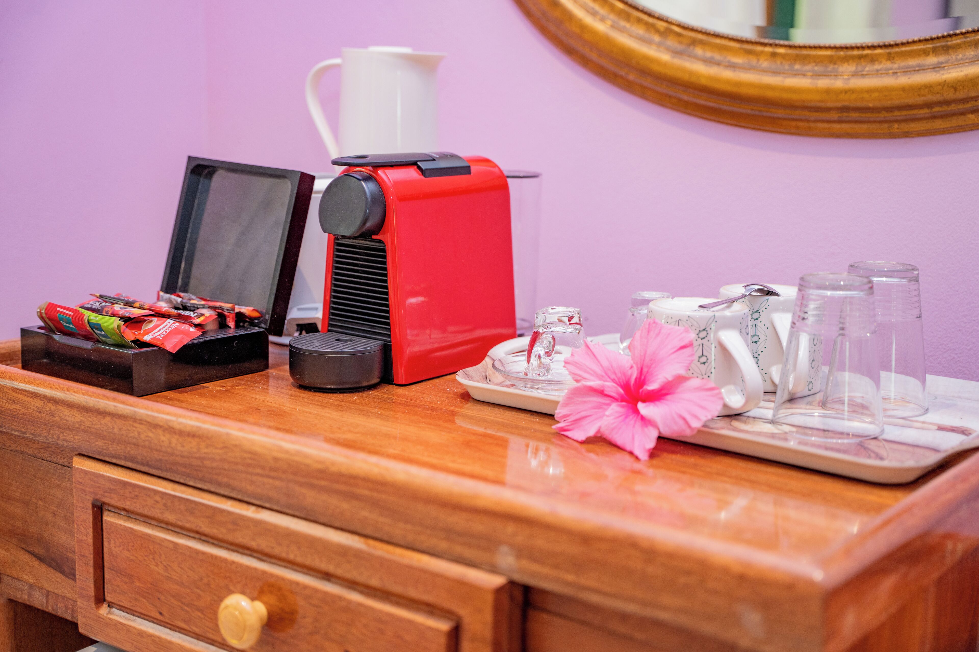 A wooden desk with a coffee maker, a tray with glasses, a flower, and a mirror.