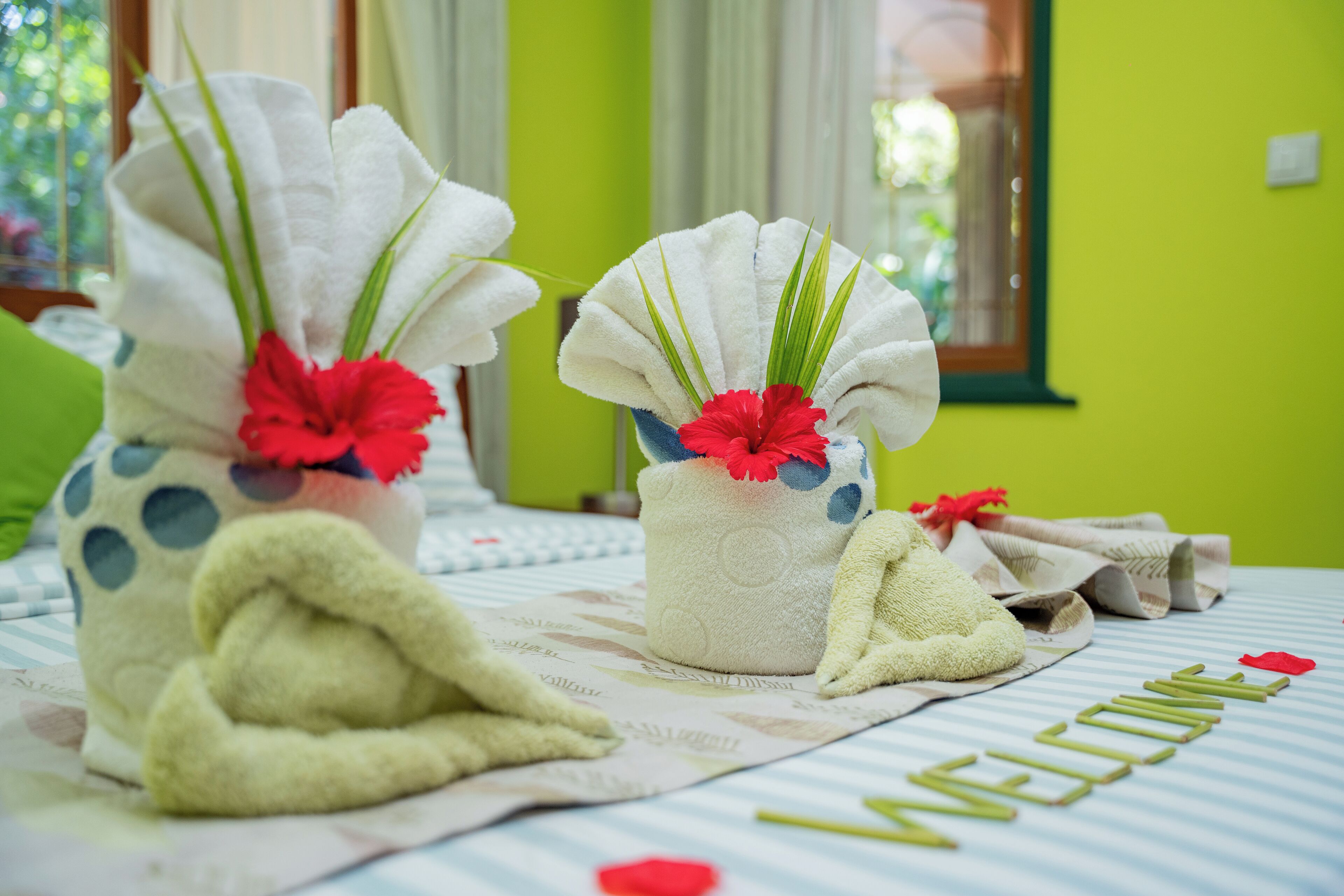Una habitación ordenada con toallas y un tapete de bienvenida sobre la cama, una pared verde y una ventana con cortinas.