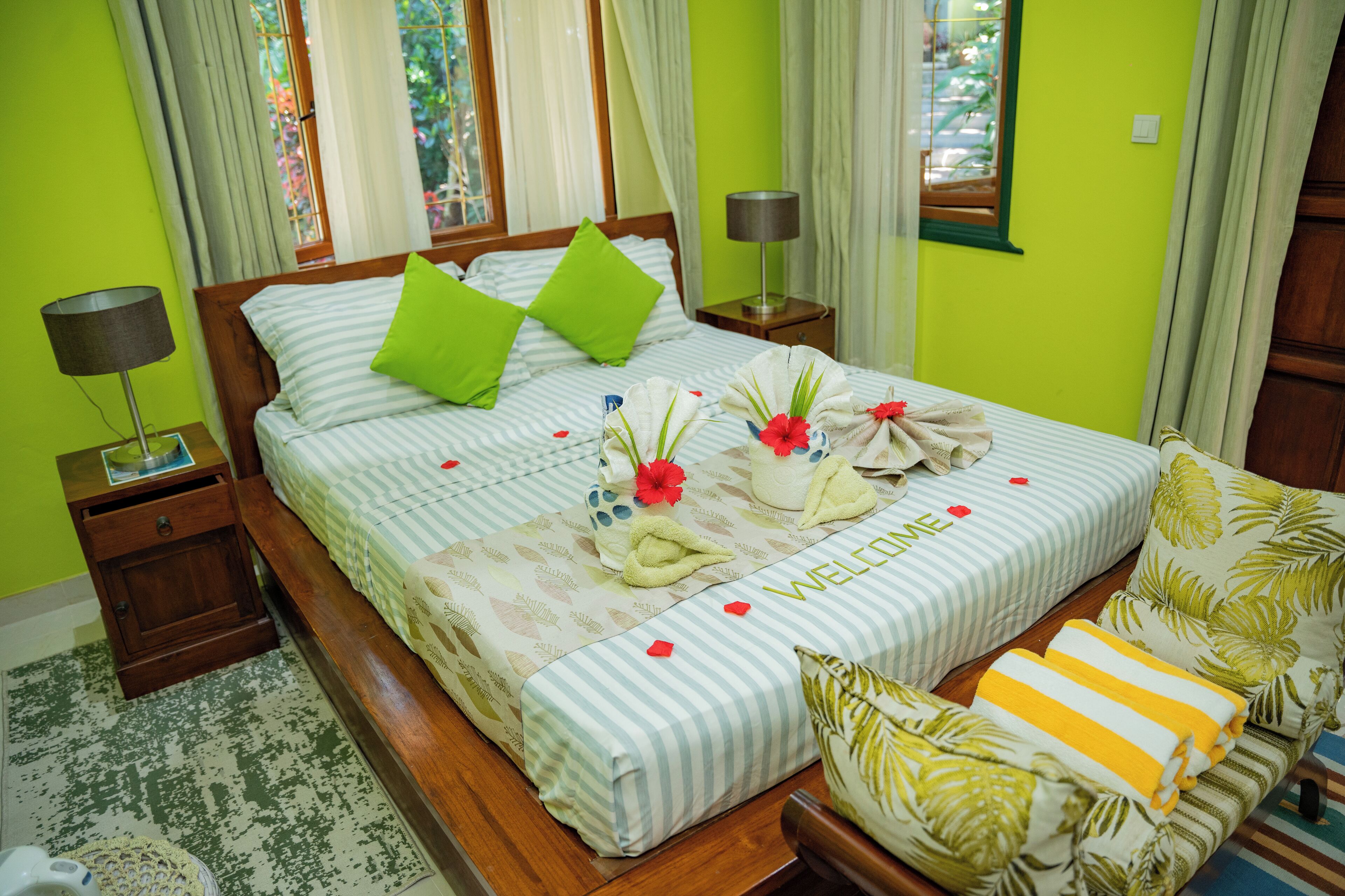 A neatly made bed with striped bedding, green pillows, and a welcome mat with flowers.