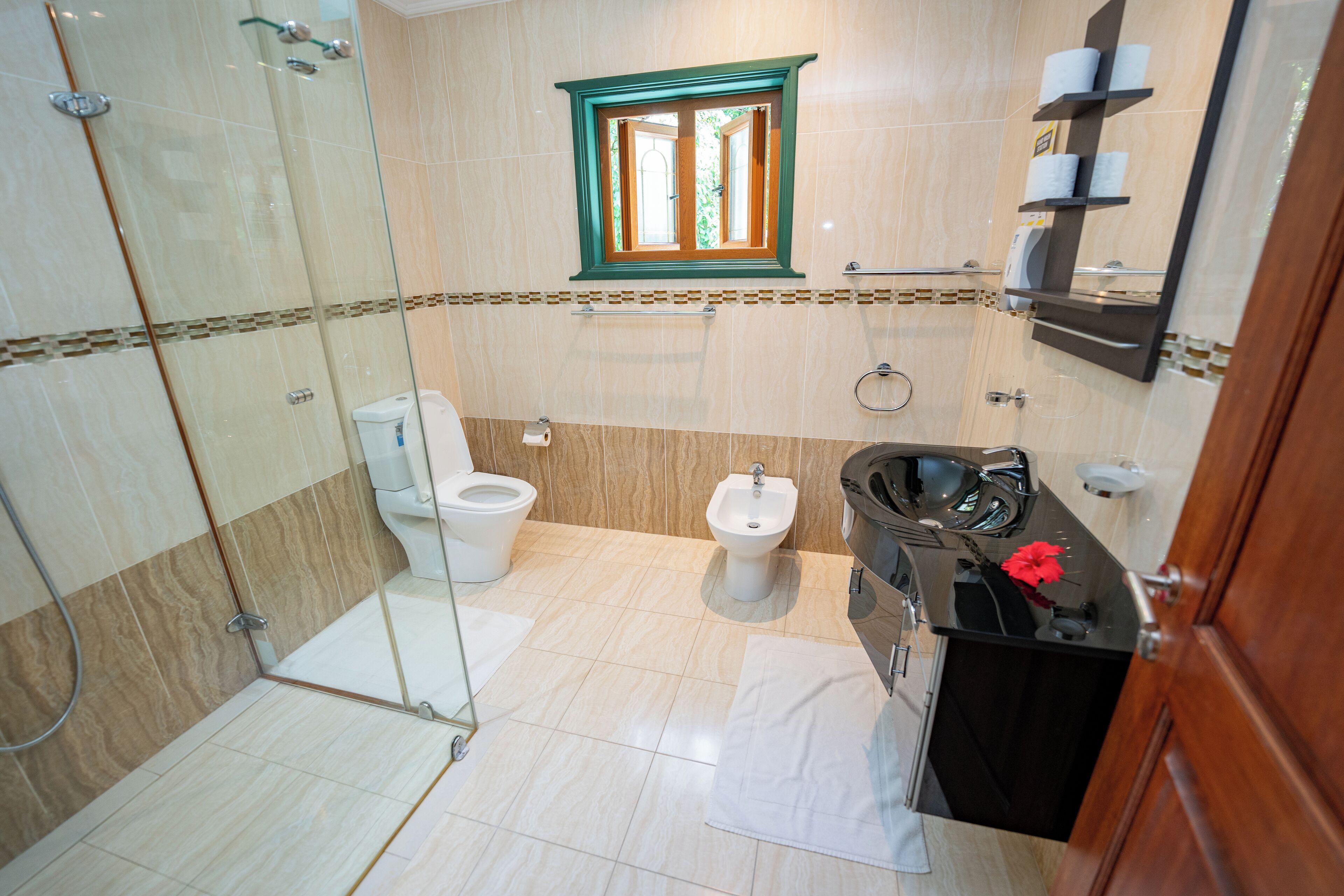 A bathroom with a glass shower enclosure, two toilets, a bidet, a vanity with a mirror, and a window with a view.